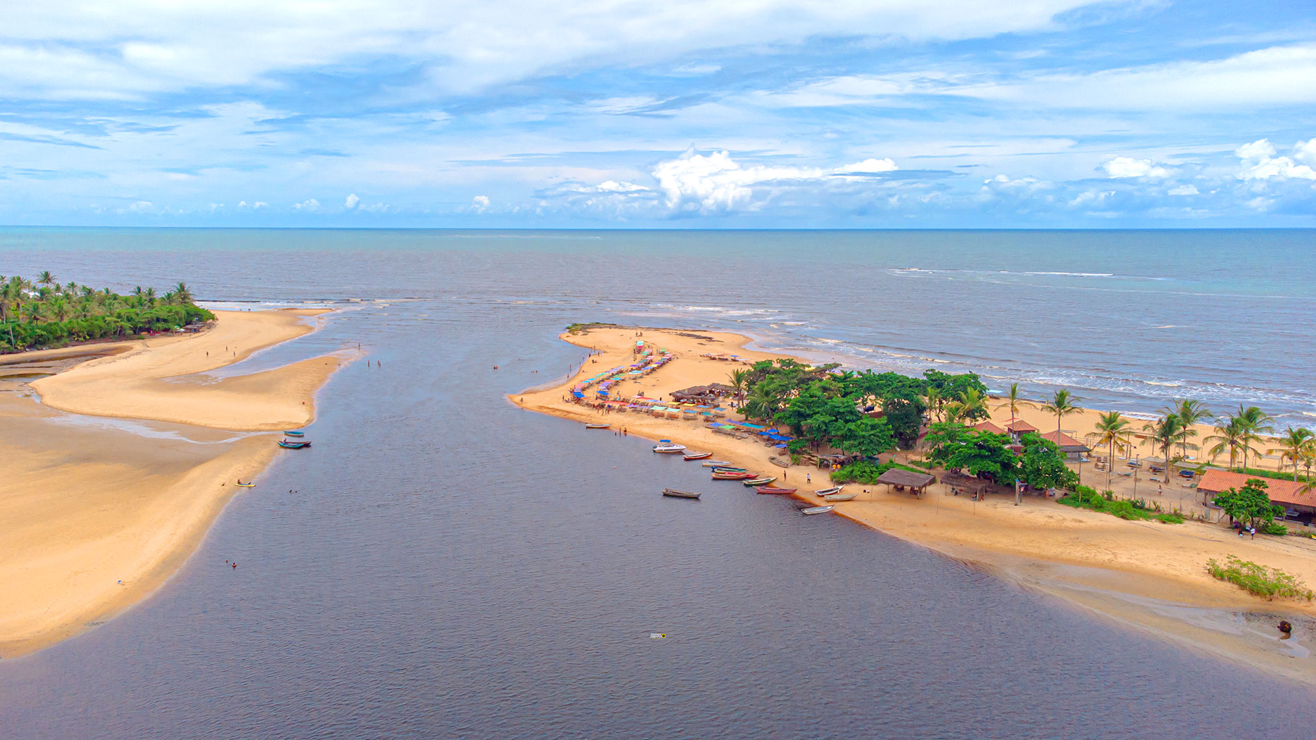 Vista aérea do encontro do Rio Caraíva com o mar, com barcos ancorados e barracas de praia coloridas