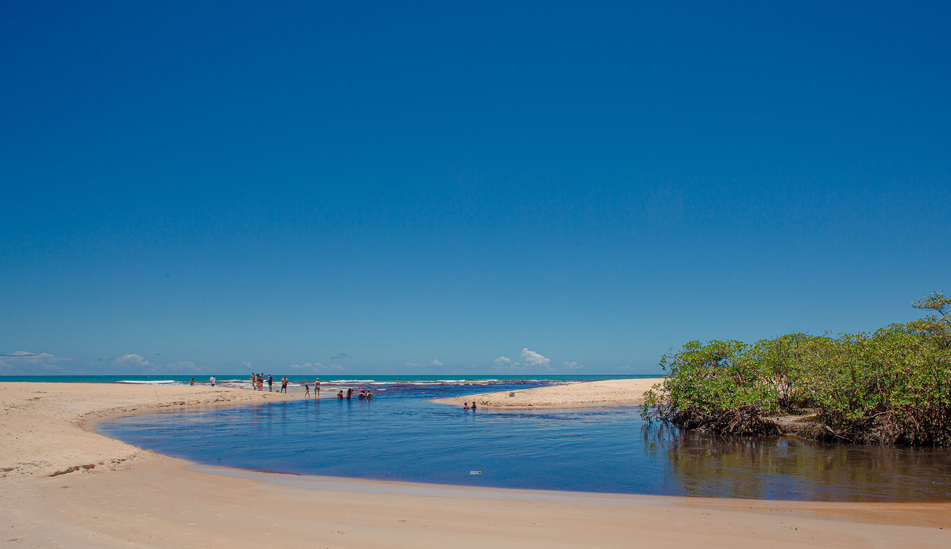 Riacho de água calma desaguando no mar em Trancoso, com pessoas tomando banho na foz